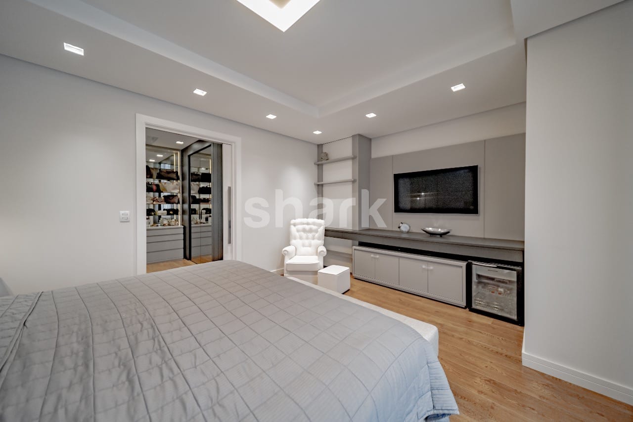 Suíte master ampla com cama king, closet iluminado ao lado e painel de TV